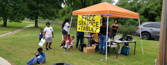 A group of individuals standing at a canopy with a banner that reads Free Service plus Tablet.