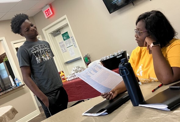 A lady sitting at a table talking to a young man who is standing.