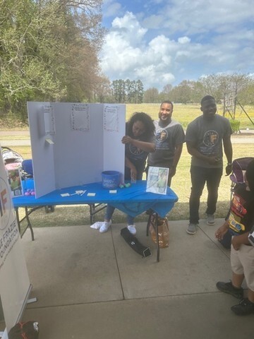 A table with a presentation board and three people standing behind it. 